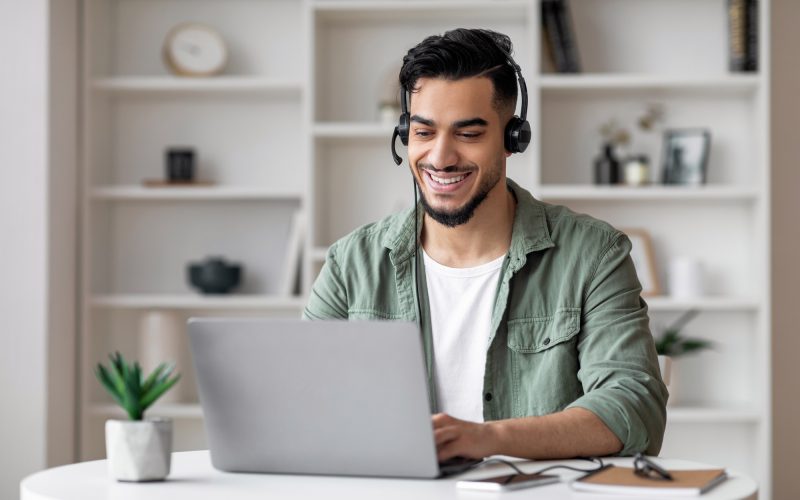 Happy handsome young islamic man with beard in headphones looks at laptop in living room interior