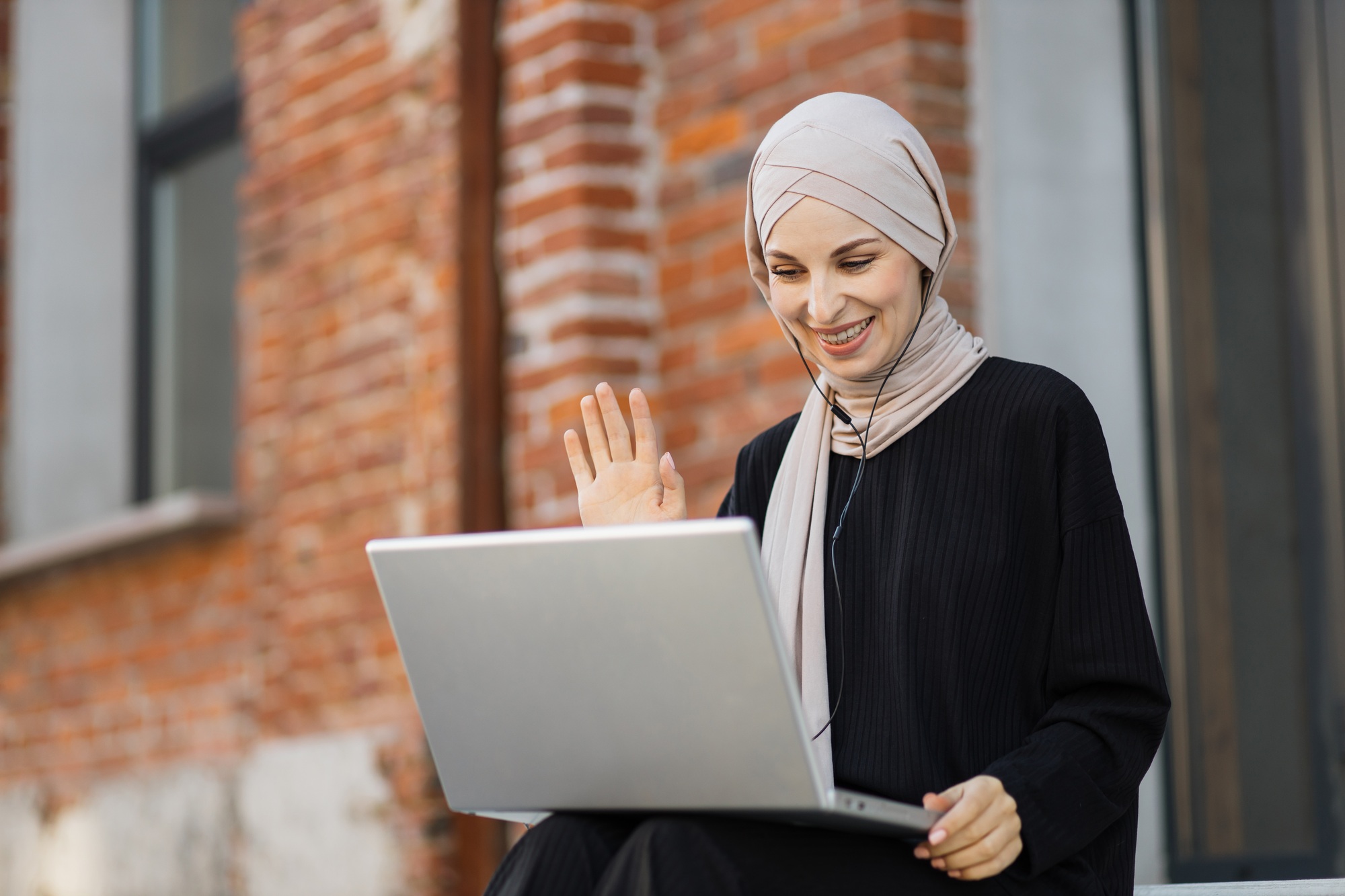 Muslim female in black clothes and hijab having online video meeting using headphones and laptop
