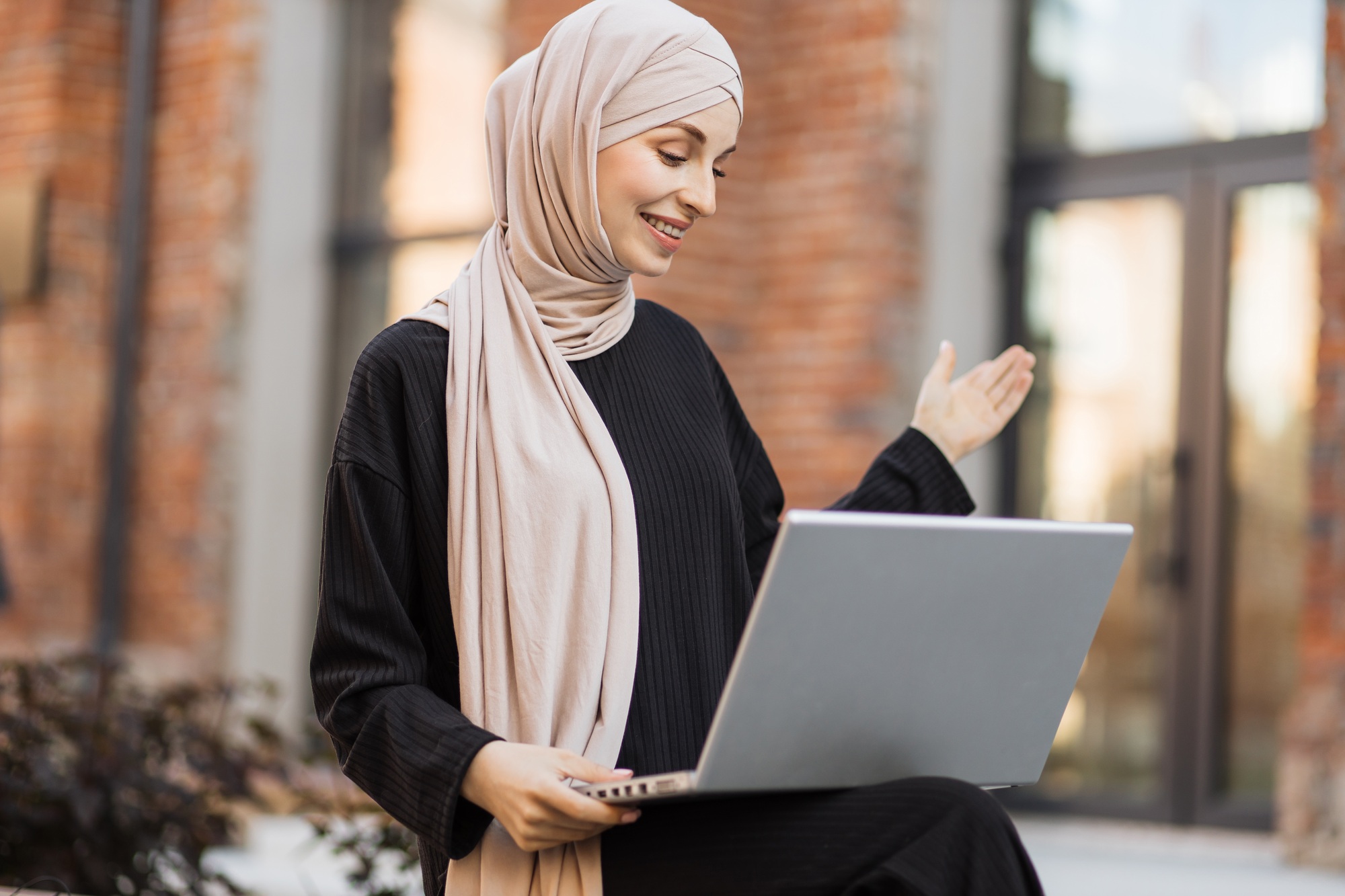 Muslim woman in hijab and black clothes sitting on bench during break talking on laptop