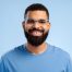Attractive, smiling, bearded African American man wearing stylish eyeglasses looking at camera standing isolated on blue background. Vision concept, treatment