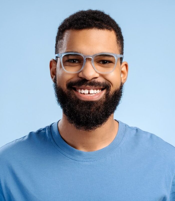 Attractive, smiling, bearded African American man wearing stylish eyeglasses looking at camera standing isolated on blue background. Vision concept, treatment