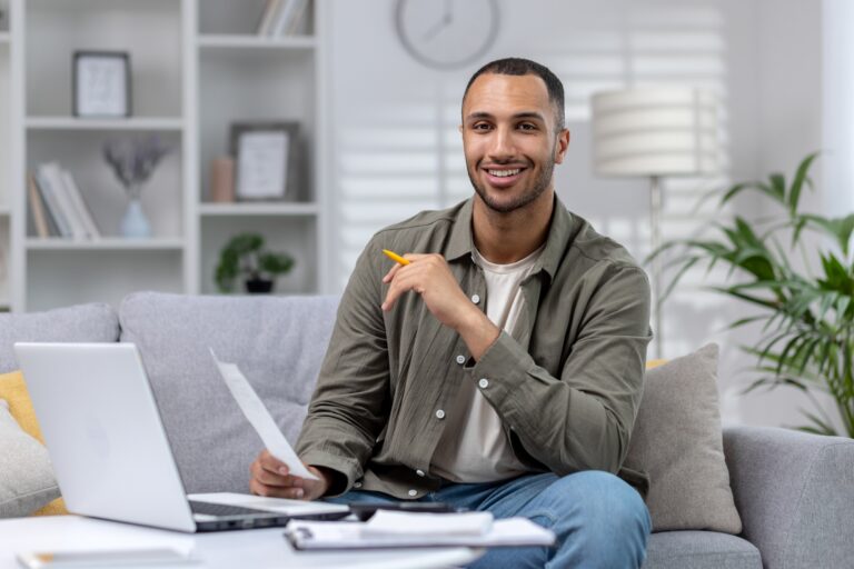 portrait-of-a-young-muslim-male-student-studying-remotely-at-home-using-a-laptop-and-smiling-at-the