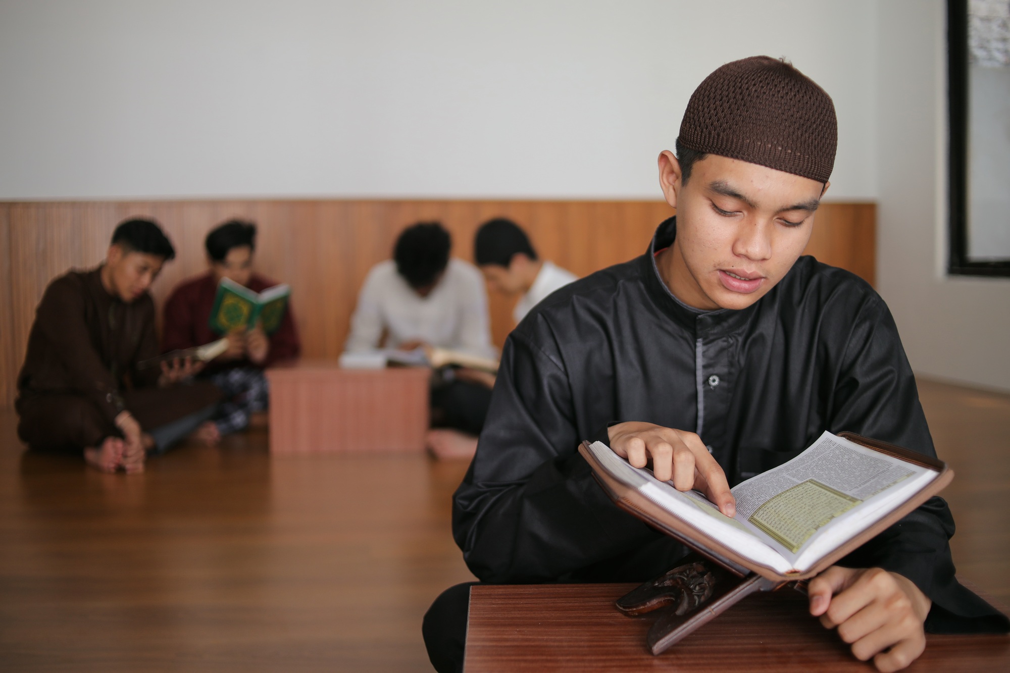 Young Muslim Boy Reading Qur'an At The Mosque Against Muslim Friends Background